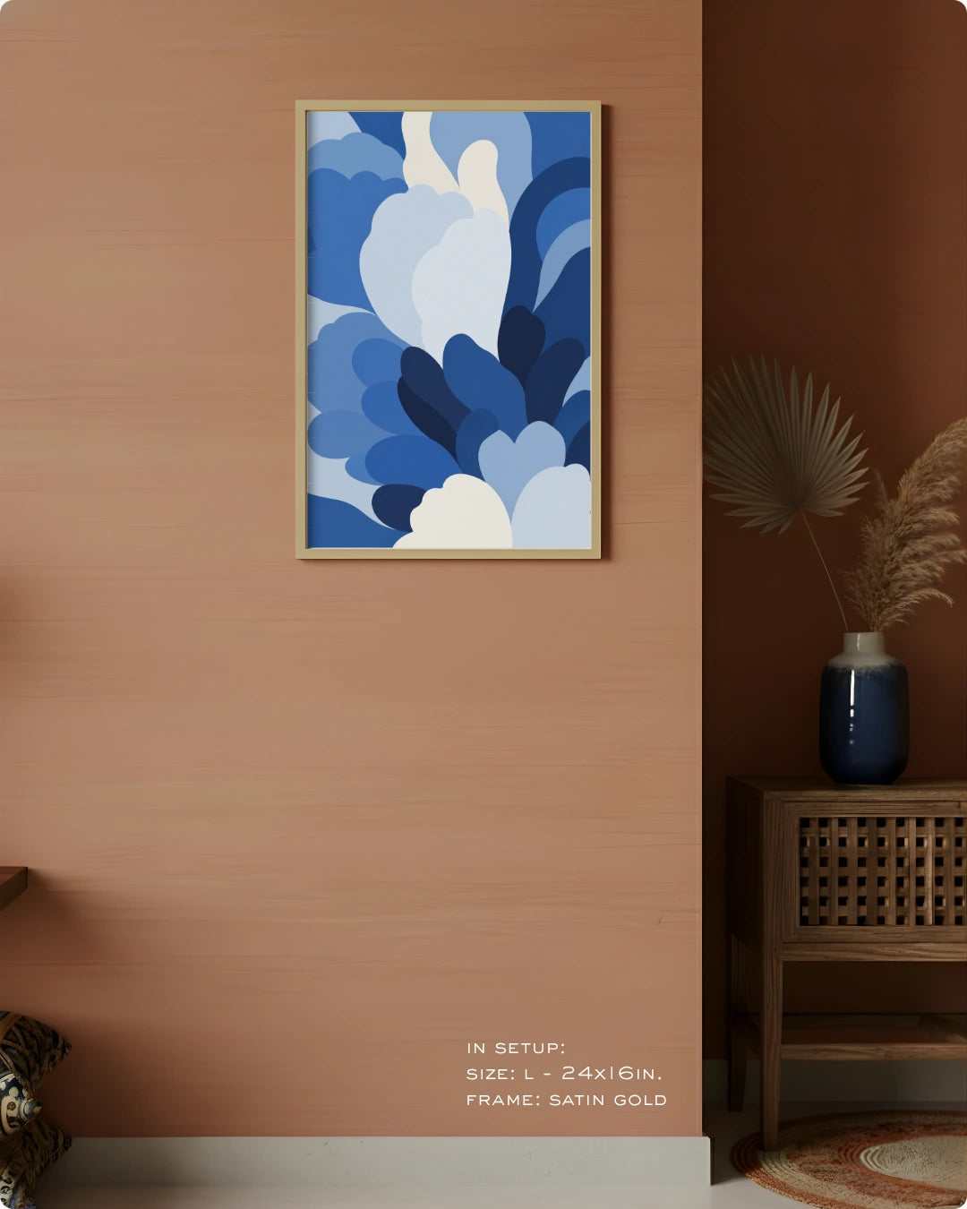 A framed abstract wall art print with layered blue and white floral-like shapes hanging on a warm peach wall in a styled interior. A small vase with dried palm leaves sits on a wooden side table to the right, and text at the bottom reads 'IN SETUP: SIZE L-24x16in.