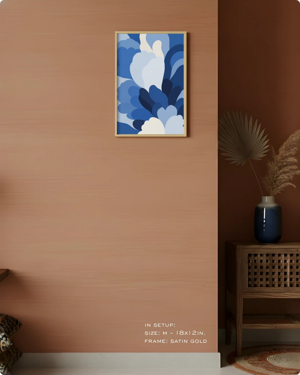 A framed abstract floral art print in shades of blue and white hangs on a warm terracotta wall in a styled interior setting with a wooden side table, vase, and dried palm leaves.