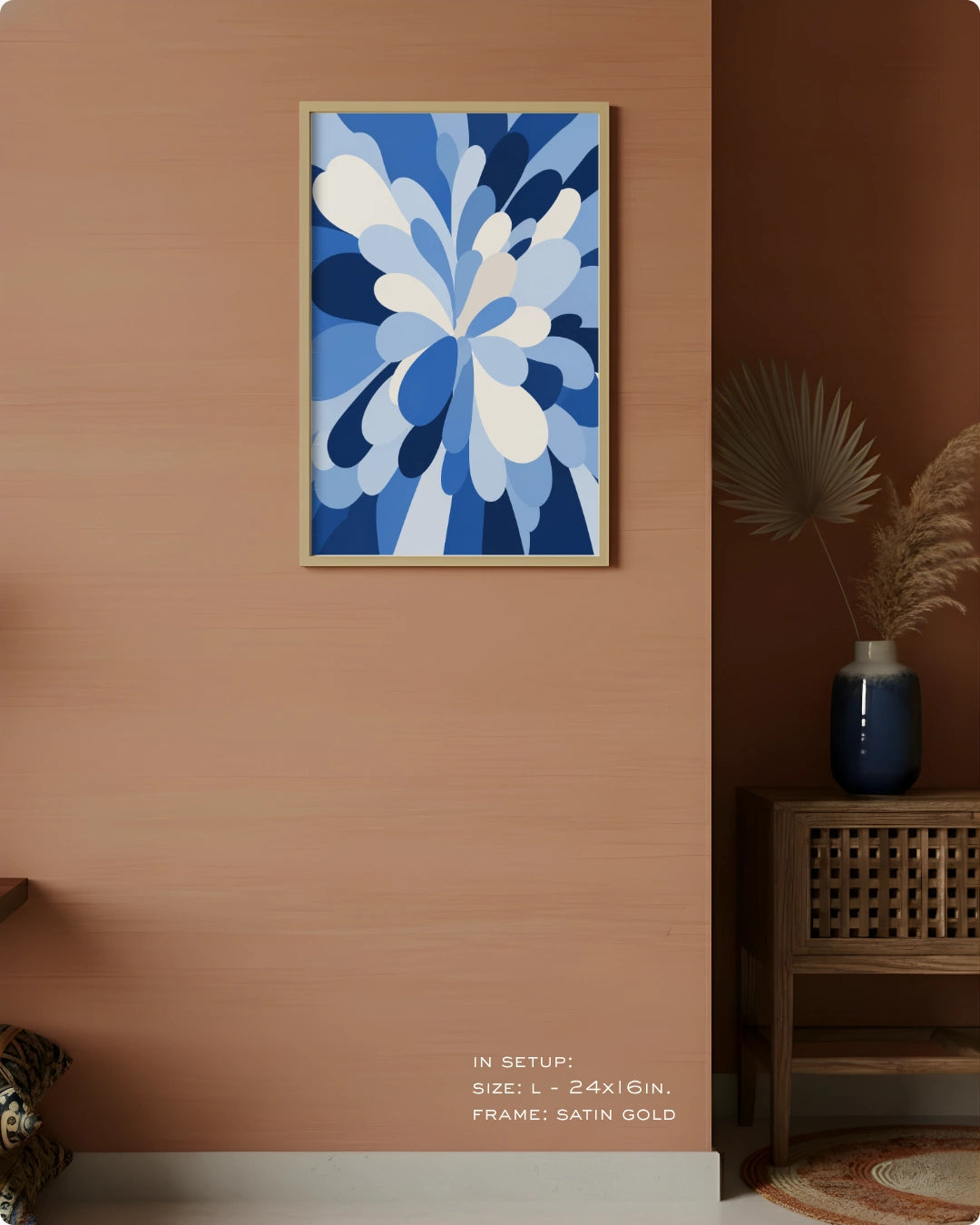 A framed abstract floral artwork in shades of blue and white hangs on a warm peach wall in a styled interior setting with a wooden side table, blue vase, dried palm leaves, and small text at the bottom indicating size and frame details.