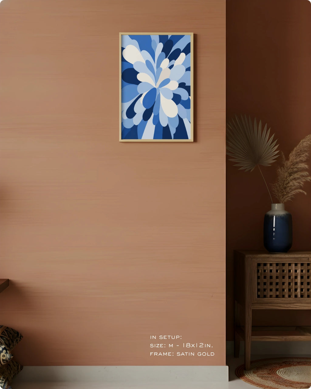 A framed abstract floral wall art print in shades of blue and white, hung on a warm terracotta-colored wall in a styled interior setting with a wooden side table, blue vase, dried palm leaves, and decorative text at the bottom.