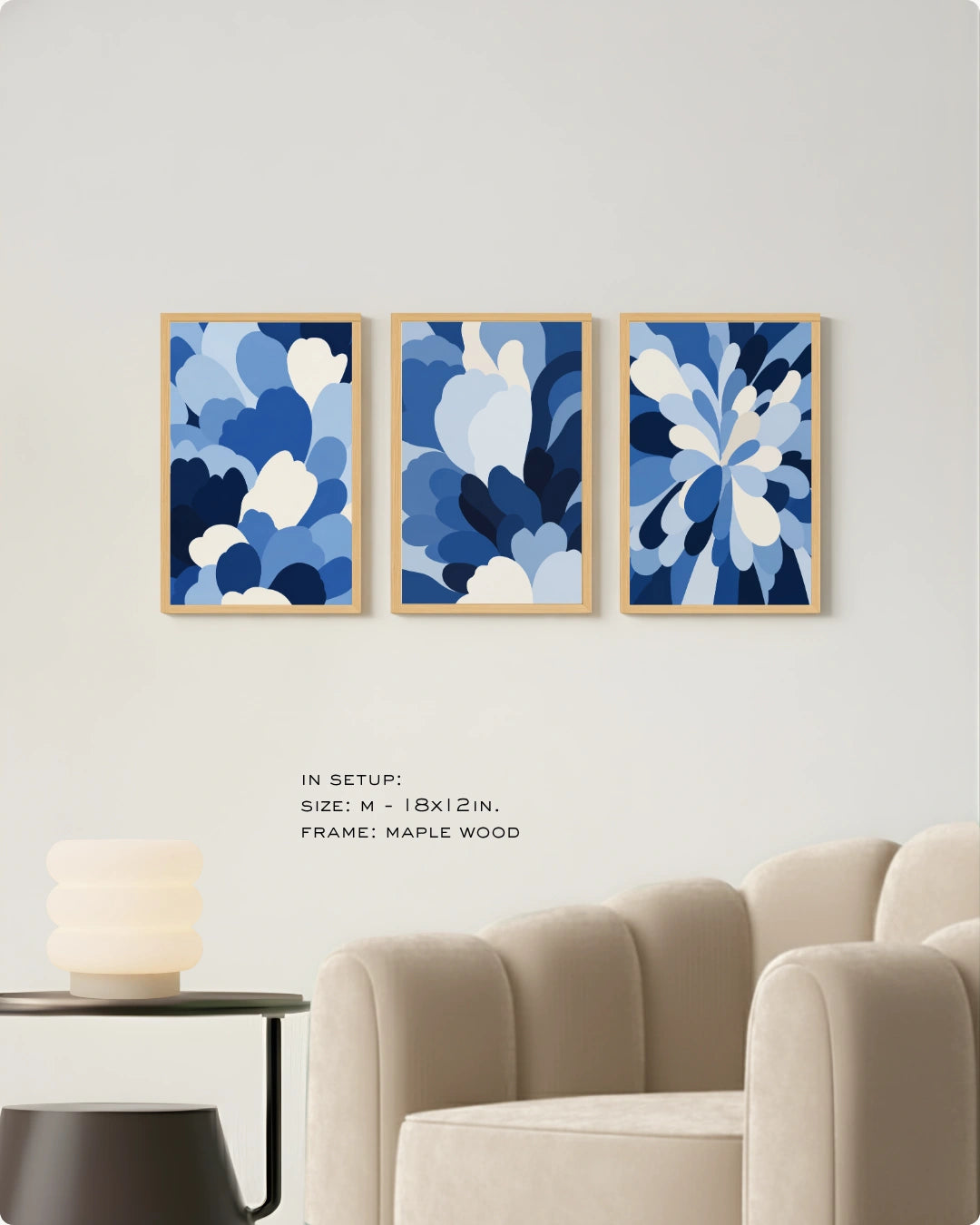 Three framed abstract blue floral art prints are displayed on a light gray wall above a beige armchair, with a small side table and lamp in the foreground. Text below reads: 'IN SETUP: SIZE: M - 18x12 IN.
