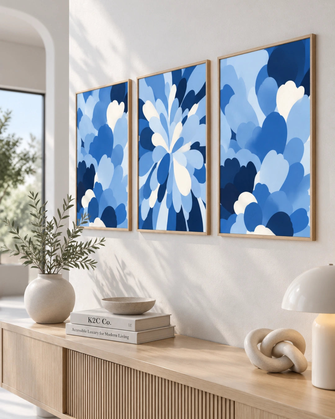 Three framed abstract canvas prints with blue floral and cloud-like patterns hang on a white wall above a light wood console table with books, a vase, and a lamp.