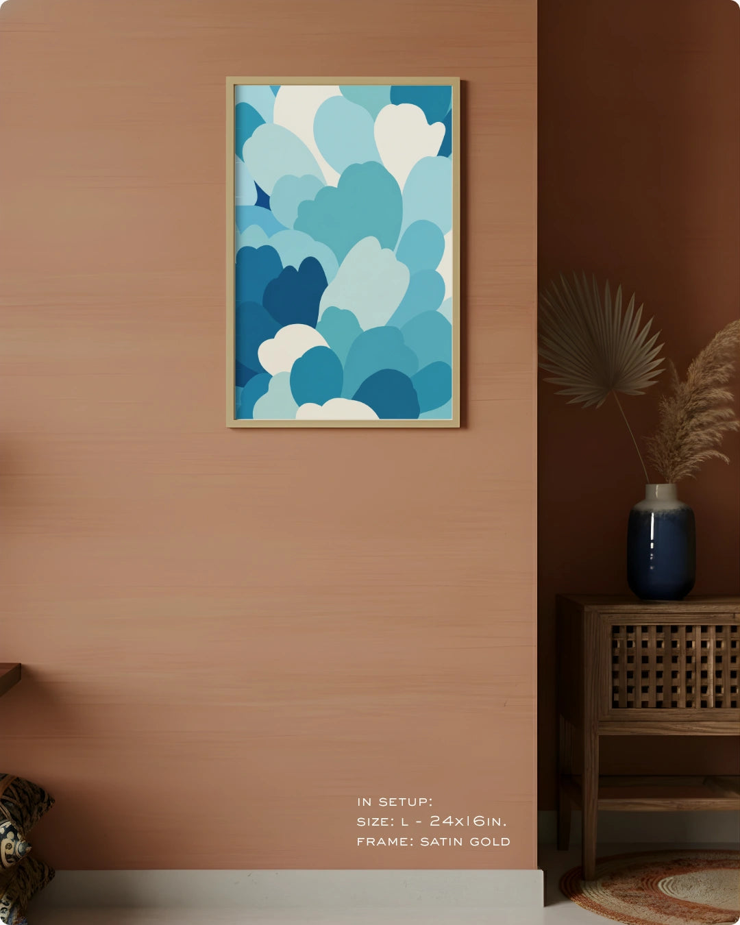 A framed abstract art print featuring layered blue and white cloud-like shapes, displayed on a warm terracotta wall in a styled interior setting with a side table and vase.