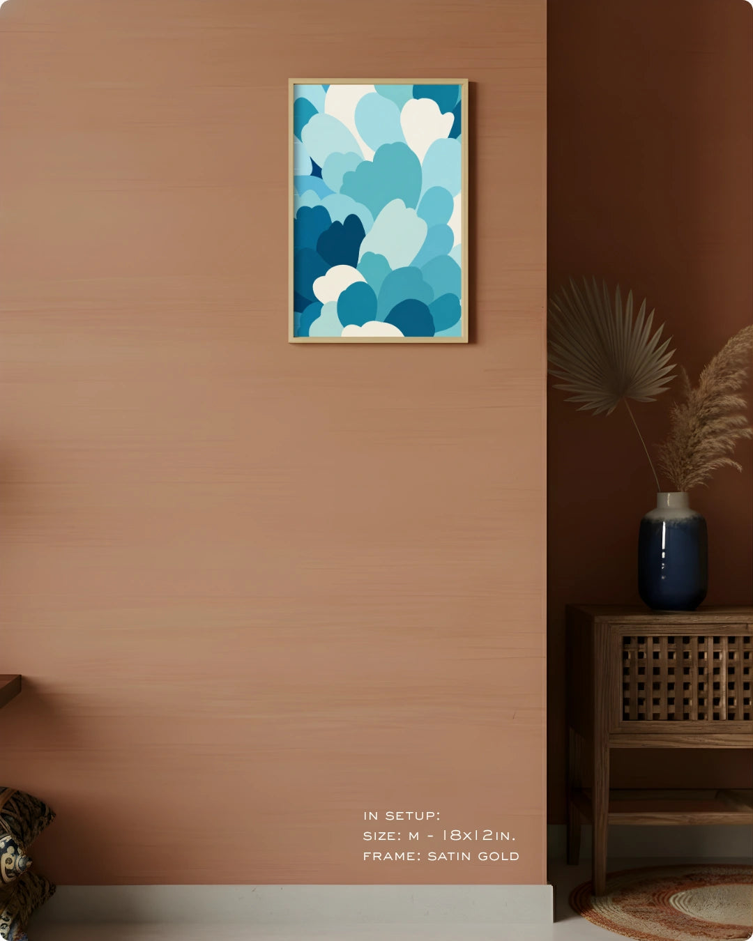 A framed abstract wall art print with layered blue and white cloud-like shapes hangs on a warm terracotta wall in a styled interior. A wooden side table with a blue vase and dried palm leaves sits to the right, and small text at the bottom reads 'IN SETUP: SIZE = 11x12 IN, FRAME: SATIN GOLD'.