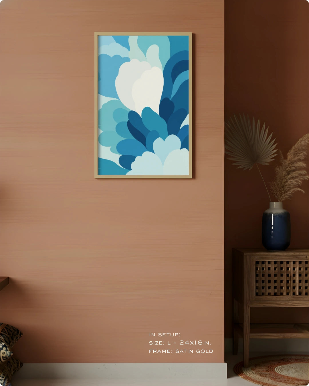 A framed abstract floral art print in shades of blue and white, hanging on a warm peach-colored wall in a styled interior setting with a wooden side table and vase.