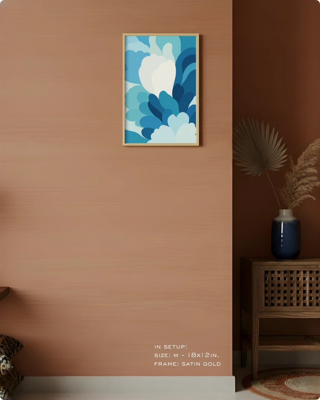 A framed abstract wall art print with layered blue and white floral-like shapes hangs on a warm terracotta wall in a styled interior. A wooden side table with a blue vase and dried palm leaves sits to the right, and small text at the bottom reads 'IN SETUP: SIZE - 1Bx2IN.