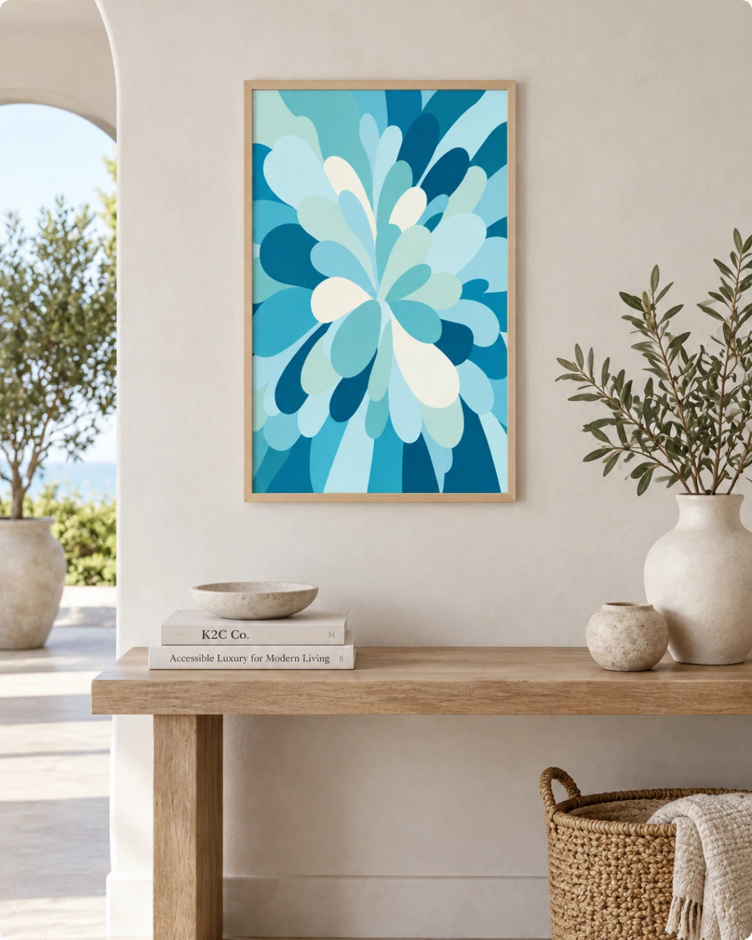A framed abstract floral art print in shades of blue and white hangs on a beige wall above a wooden console table styled with vases, books, and a basket.