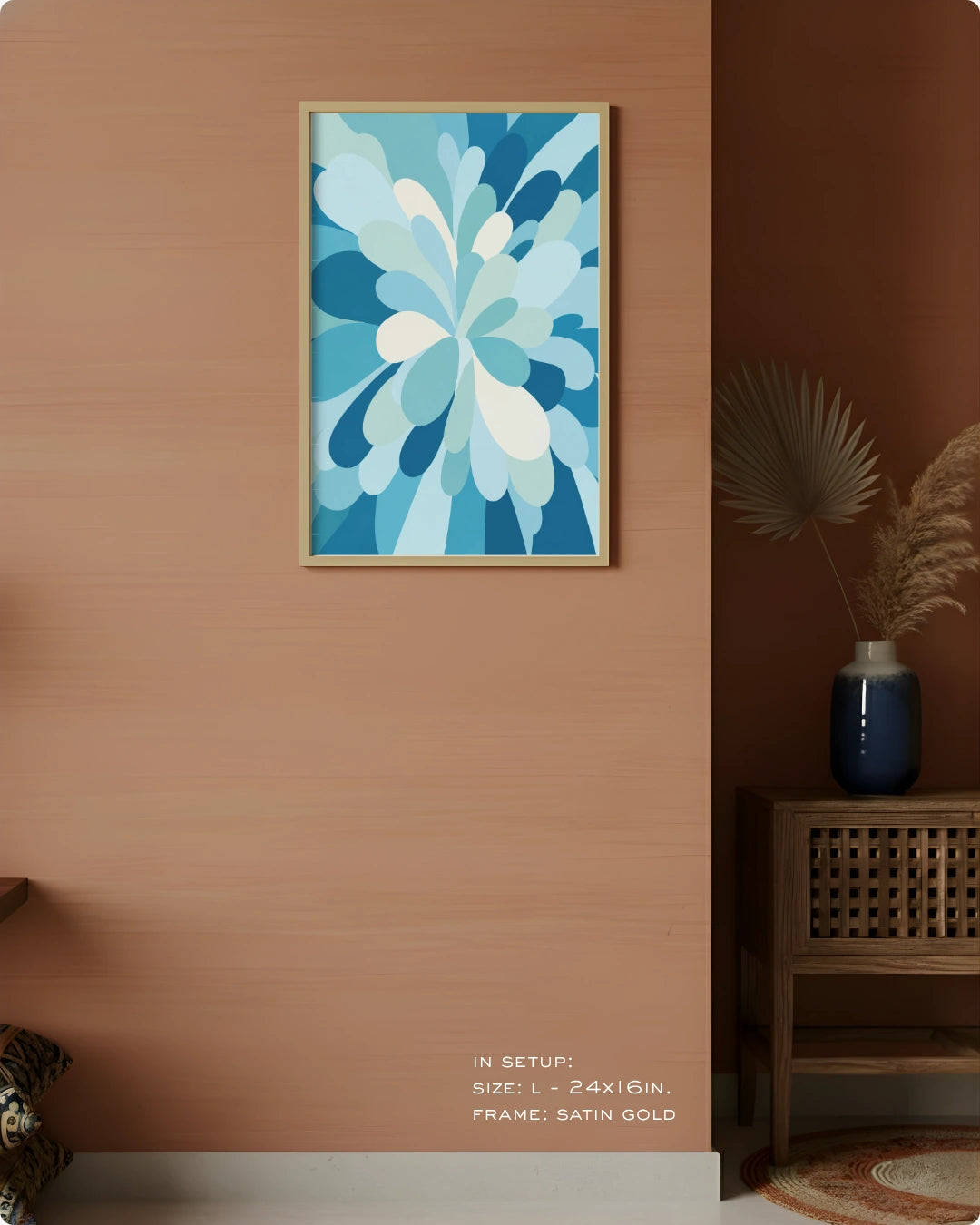 A framed abstract floral art print in shades of blue and white, hanging on a warm peach-colored wall in a styled interior setting with a wooden side table, blue vase, and dried palm leaves.
