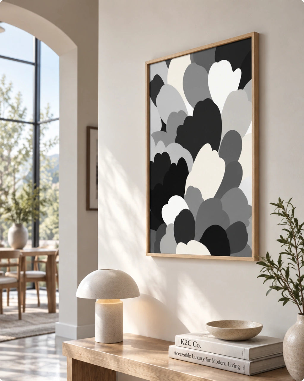 A framed abstract wall art print with overlapping cloud-like shapes in black, gray, and white, hanging on a beige wall in a modern, sunlit living space with a console table, lamp, and books.