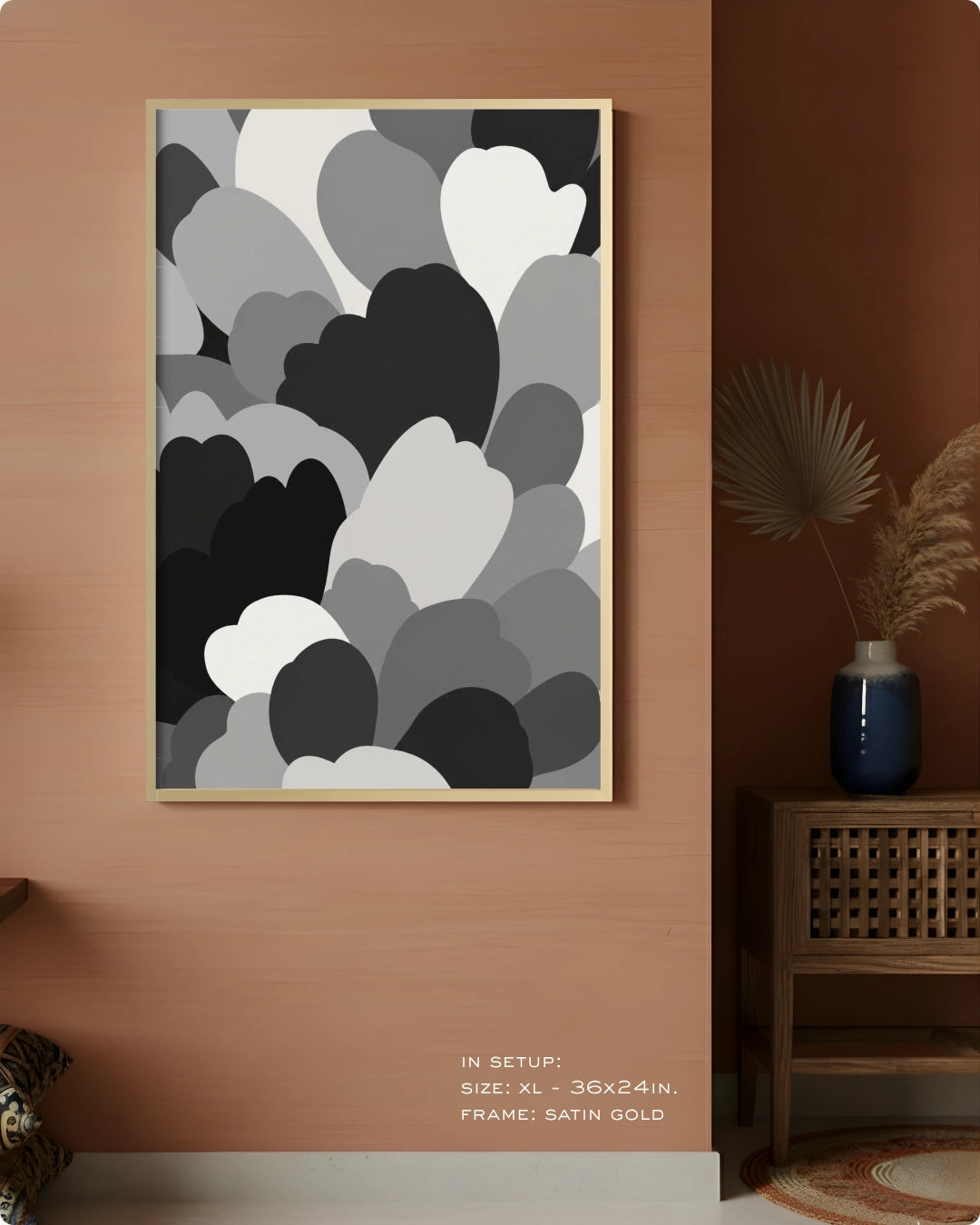 A framed abstract wall art print with layered gray, black, white, and charcoal cloud-like shapes, displayed on a warm terracotta wall in a styled interior setting with a side table and vase.