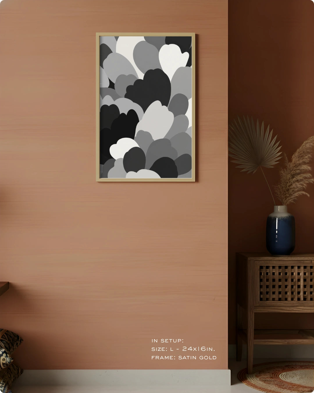 A framed abstract wall art print with layered black, gray, and white cloud-like shapes hanging on a warm peach wall in a styled interior. A wooden side table with a blue vase and dried palm leaves sits to the right, and small text at the bottom reads 'IN SETUP: SIZE L = 24x16in.