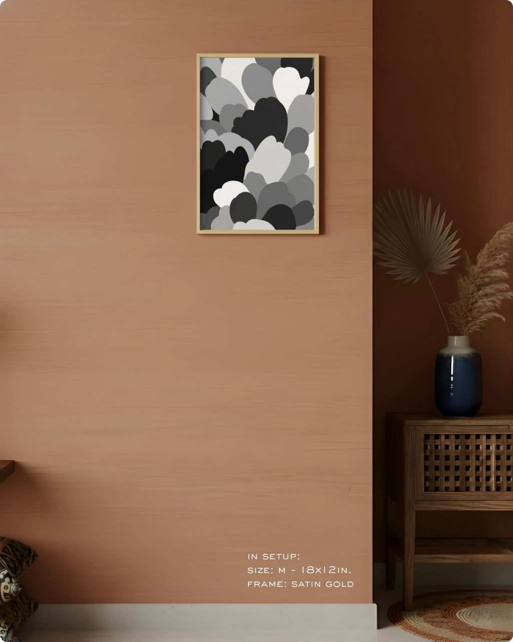 A framed abstract wall art print with overlapping cloud-like shapes in black, gray, and white, hung on a warm terracotta wall in a styled interior setting with a wooden side table, blue vase, dried palm leaves, and small text at the bottom indicating size and frame details.