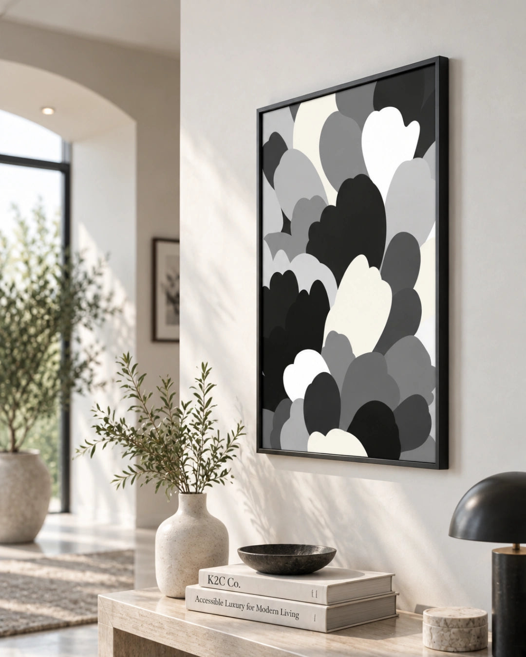 A framed abstract wall art print with overlapping cloud-like shapes in black, gray, and white, hanging on a light wall in a modern interior setting with a console table, vase, books, and lamp.