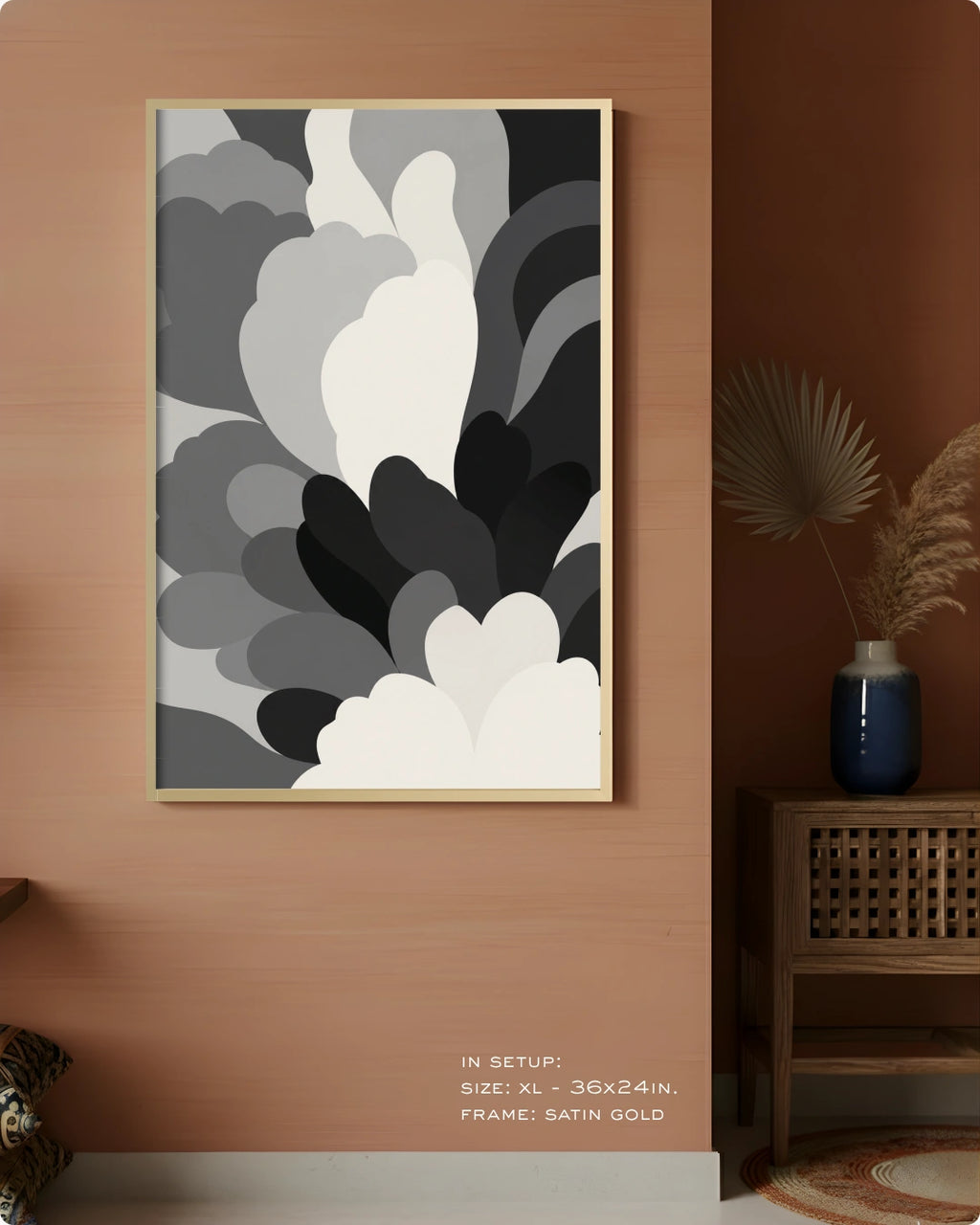 A framed abstract wall art print with layered gray, white, and black organic shapes hanging on a warm terracotta wall in a styled interior. The frame is satin gold.