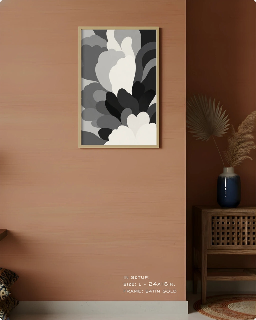 A framed abstract wall art print hangs on a warm peach-colored wall in a styled interior. The artwork features layered organic shapes in white, gray, and black, enclosed in a thin satin gold frame.