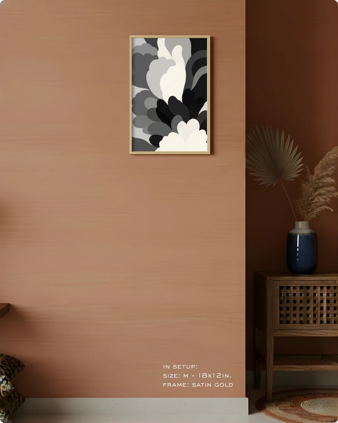A framed abstract wall art print with black, gray, white, and brown organic shapes, hung on a terracotta-colored wall in a styled interior setting with a wooden side table, blue vase, and dried palm leaves.