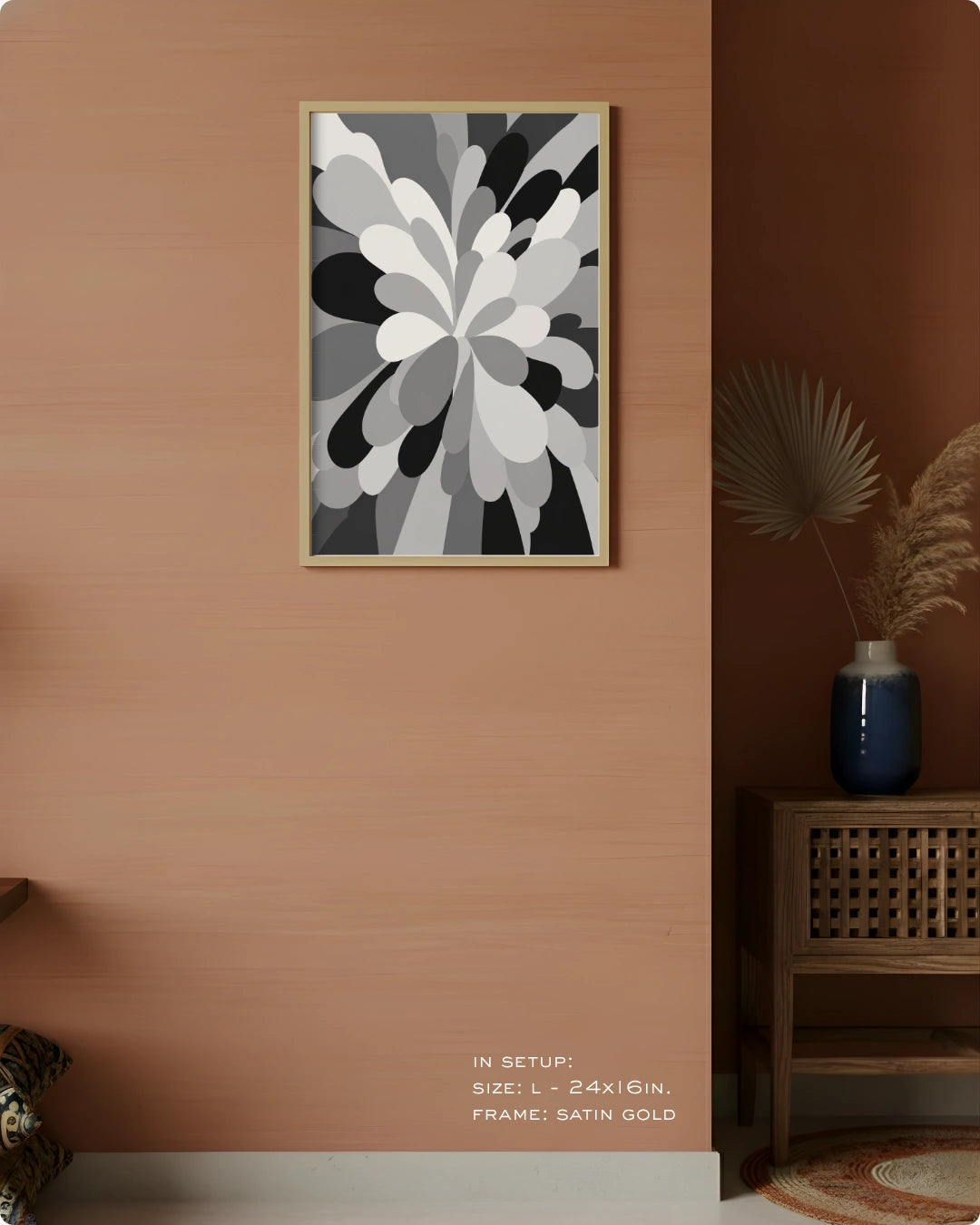 A framed abstract floral wall art print in black, white, and gray tones hanging on a warm peach-pink wall in a styled interior setting with a wooden side table, blue vase, dried palm leaves, and a rug.