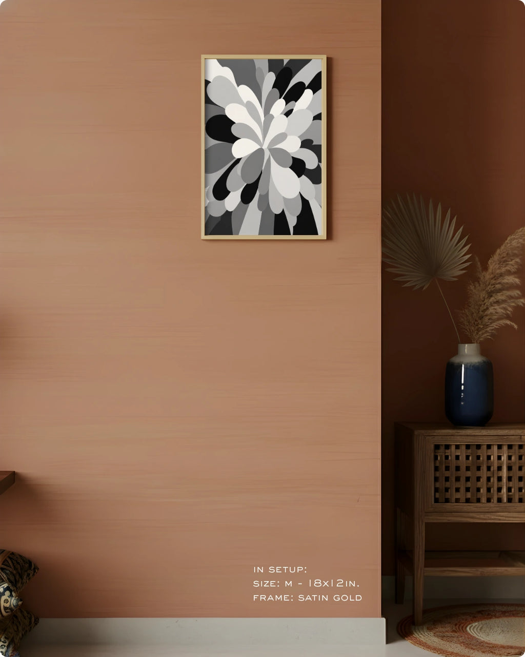A framed abstract floral wall art print in black, white, and gray hangs on a warm terracotta-colored wall in a styled interior setting with a wooden side table, blue vase, dried palm leaves, and a small rug.
