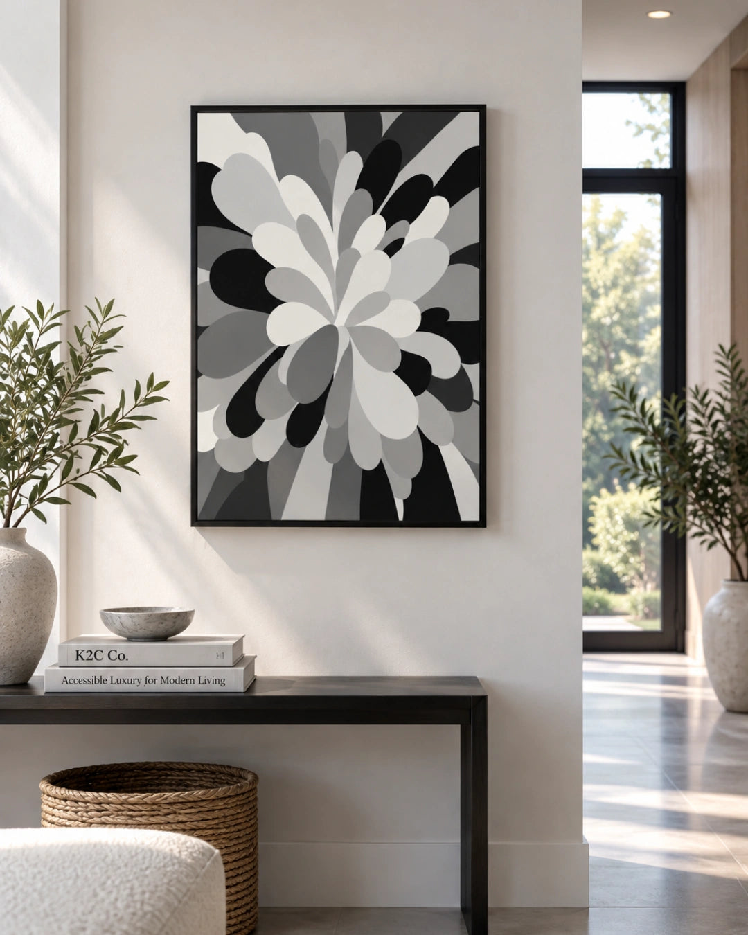 A framed abstract floral wall art print in black, gray, and white hangs on a light beige wall above a dark console table in a modern, sunlit interior.