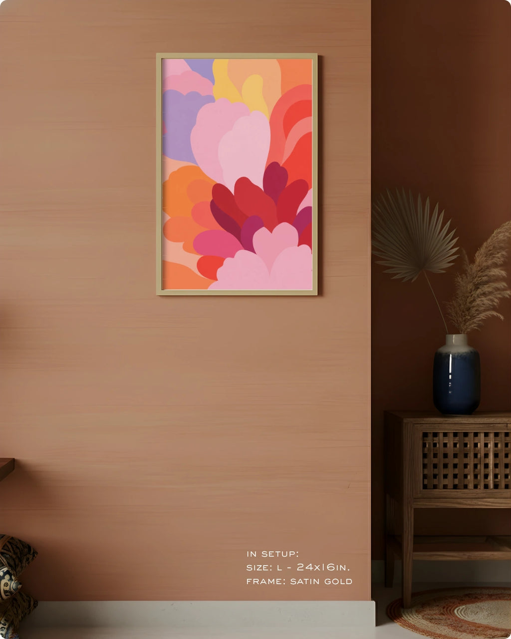 A framed abstract floral wall art print hangs on a warm peach-brown wall in a styled interior. The artwork features layered, overlapping flower-like shapes in pink, orange, red, purple, and yellow tones, set in a thin satin gold frame.