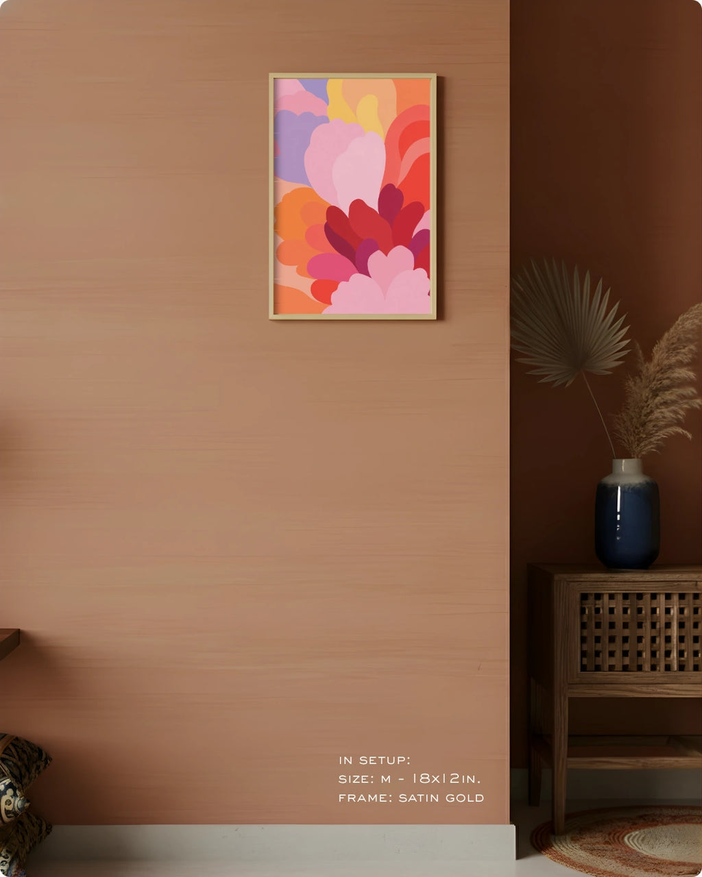 A framed abstract floral art print with overlapping pink, orange, purple, and red shapes hangs on a warm terracotta wall in a styled interior. The frame appears satin gold.