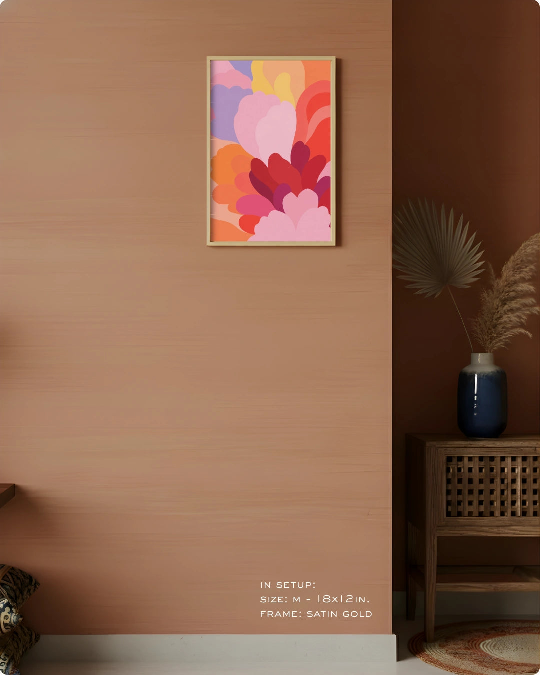 A framed abstract floral art print with overlapping pink, orange, purple, and red shapes hangs on a warm terracotta wall in a styled interior. The frame appears satin gold.