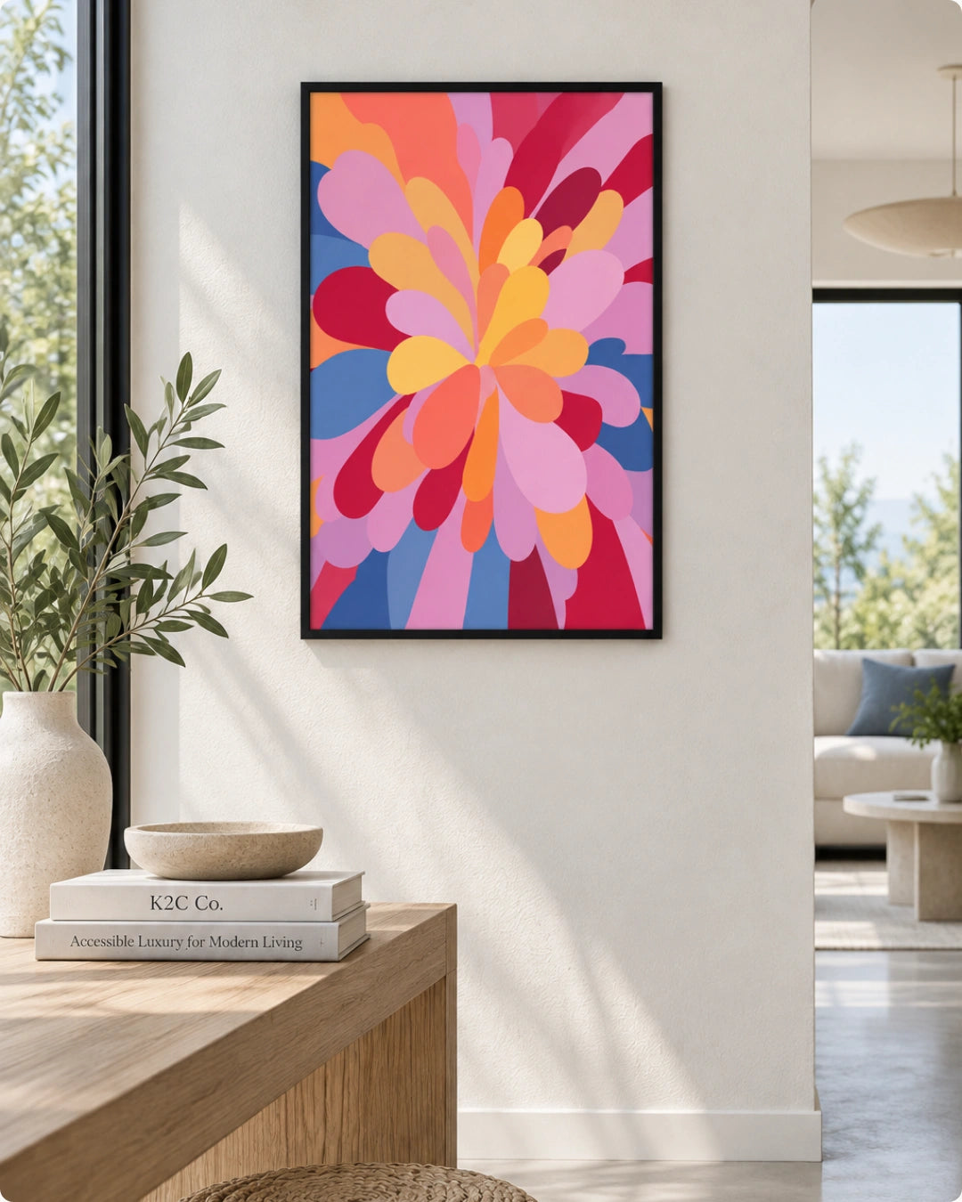 A framed abstract floral art print with bold pink, orange, yellow, and blue petal-like shapes hanging on a white wall in a modern living space. The print is shown in a black frame, styled above a wooden console table with books and a vase.