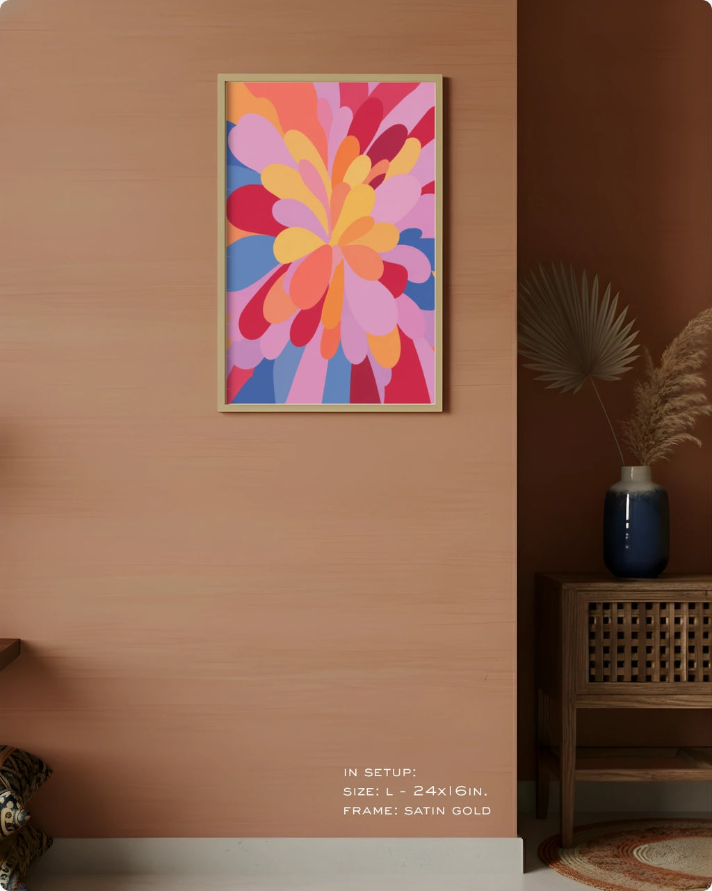 A framed abstract floral art print with vibrant pink, orange, yellow, and blue petal-like shapes hangs on a warm peach wall in a styled interior setting. The frame is satin gold.