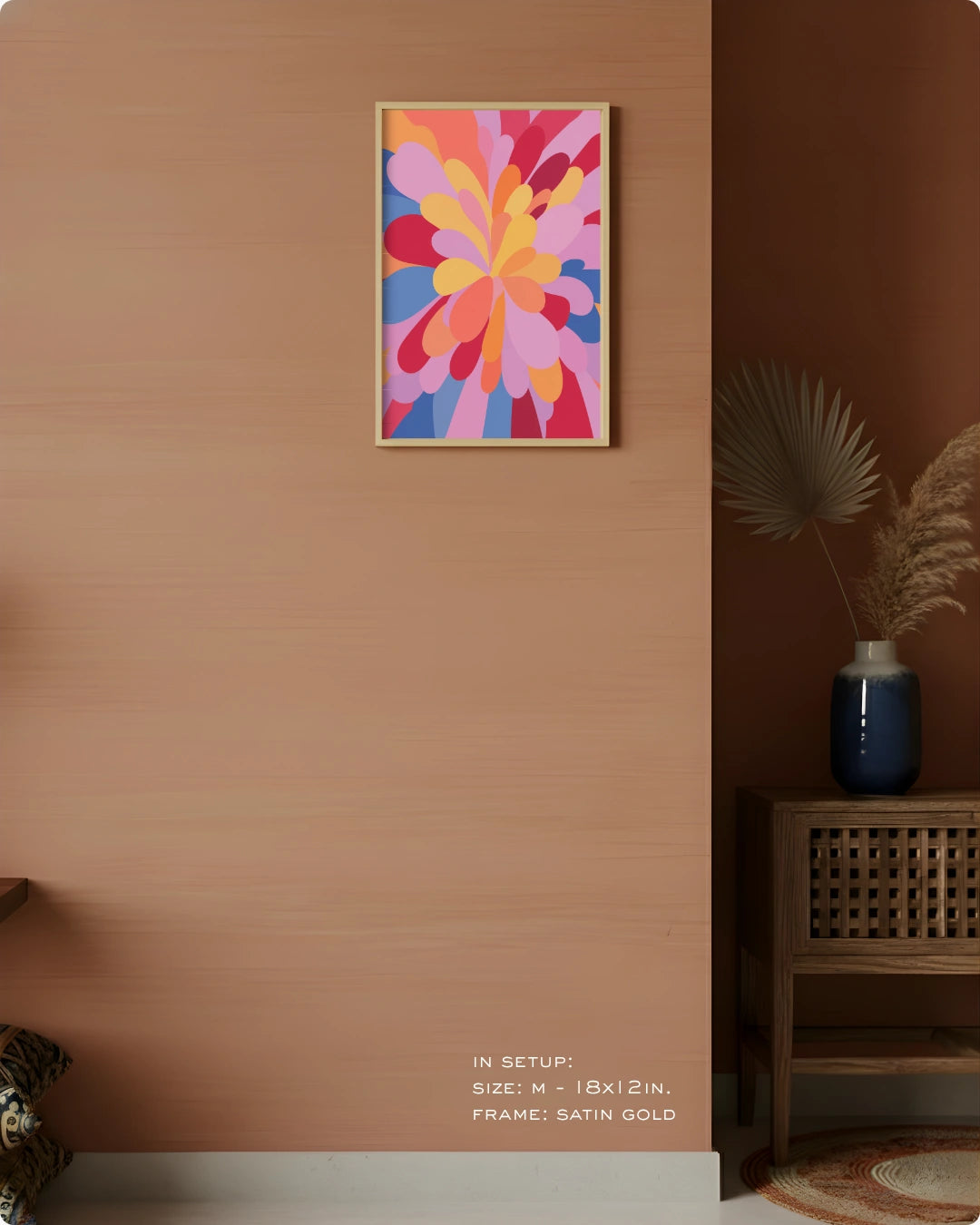 A colorful abstract floral wall art print with pink, yellow, orange, and blue shapes, framed in satin gold, hanging on a warm beige wall in a styled interior setting.