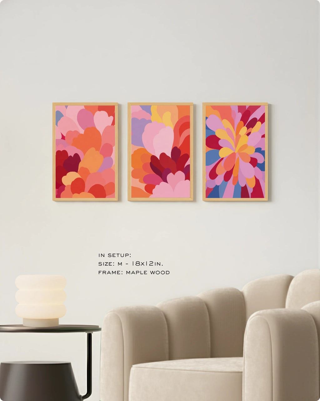Three framed abstract floral art prints in warm pink, orange, red, and purple tones are displayed on a white wall above a beige armchair, with small text noting the setup size and maple wood frame.