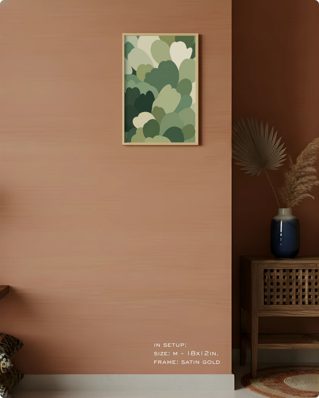 A framed abstract wall art print with layered green and cream leaf-like shapes hangs on a warm terracotta wall in a styled interior. A wooden side table with a blue vase and dried palm leaves sits to the right, and small text at the bottom reads 'IN SETUP: SIZE - 18x12 IN.