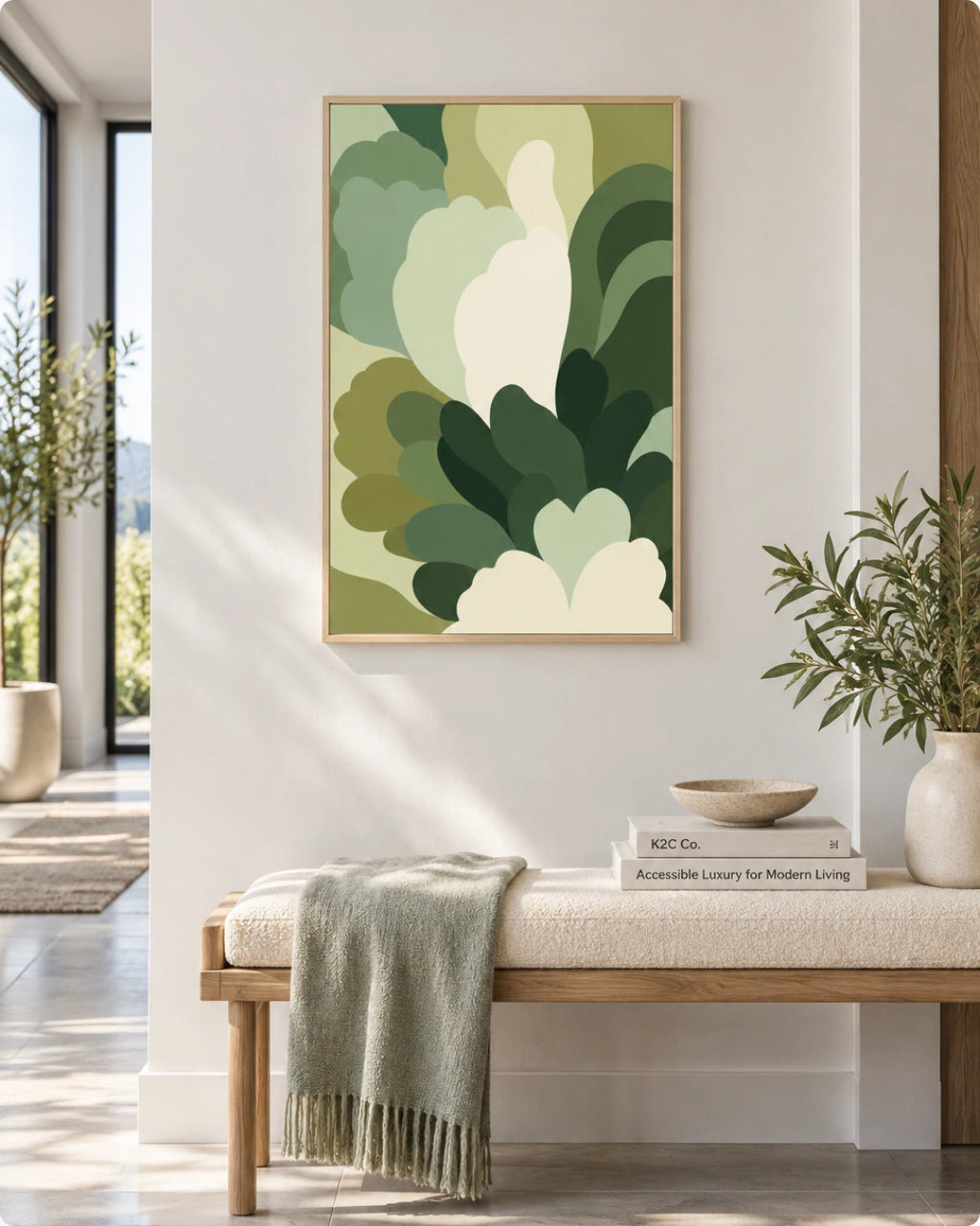 A framed abstract botanical art print with layered green and cream leaf-like shapes hangs on a white wall above a light wood bench with a throw blanket and books in a bright, modern interior.