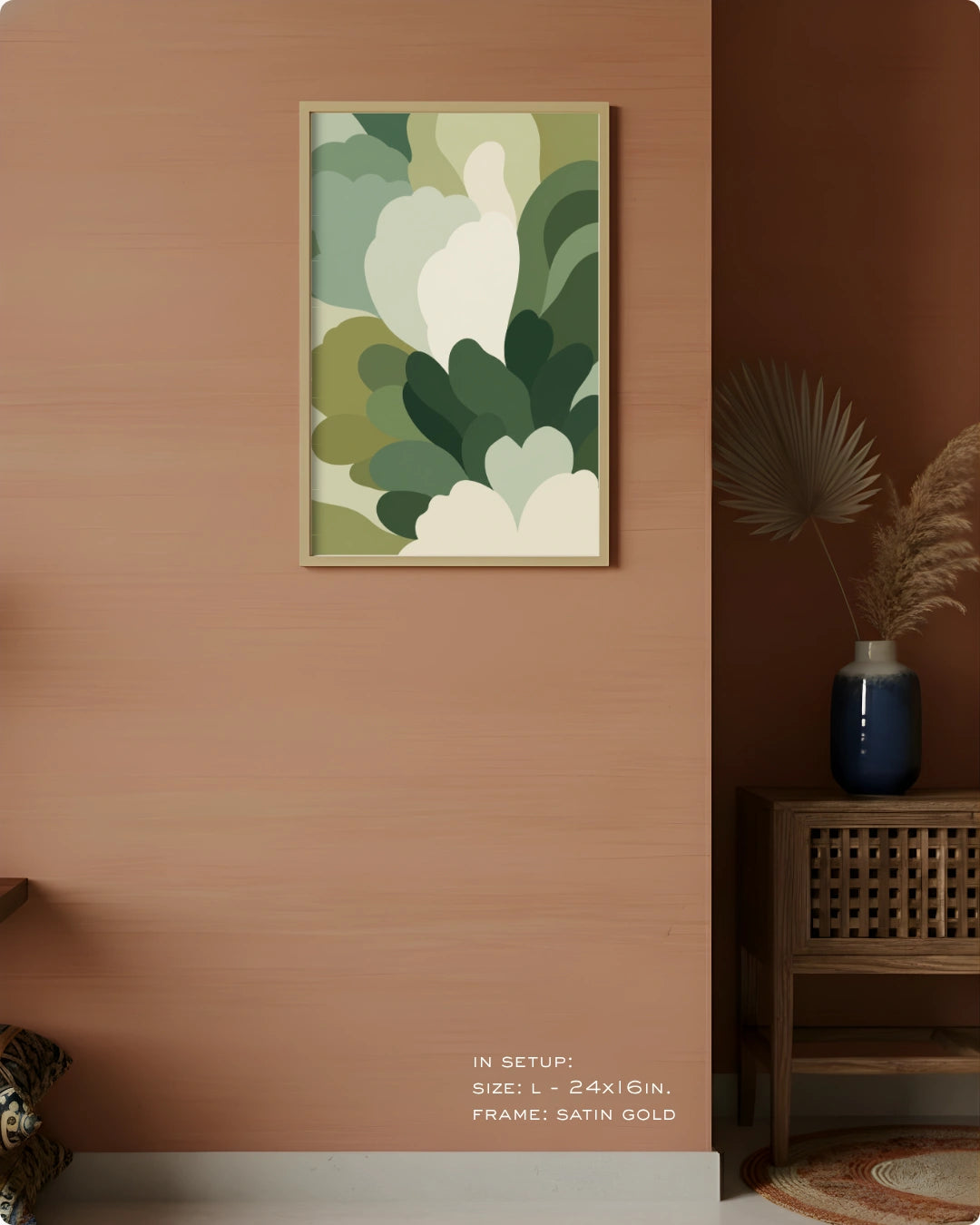 A framed abstract botanical art print with layered green and white leaf-like shapes, displayed on a warm peach-pink wall in a styled interior setting with a wooden side table, blue vase, dried palm leaves, and small text at the bottom indicating size and frame details.