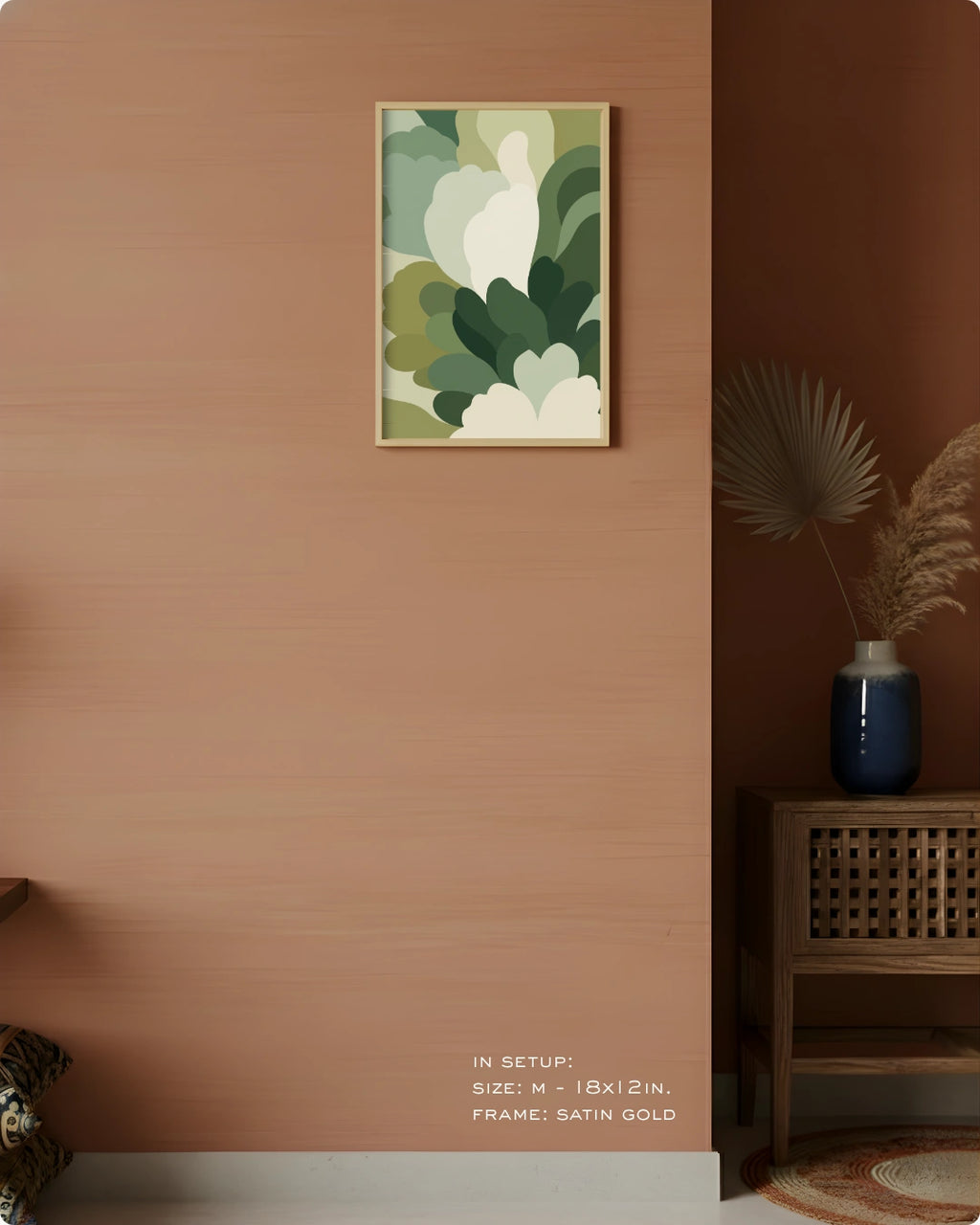 A framed abstract botanical art print with green, beige, and white organic shapes hangs on a warm terracotta wall in a styled interior. A dark wood side table with a blue vase and dried palm leaves sits to the right.