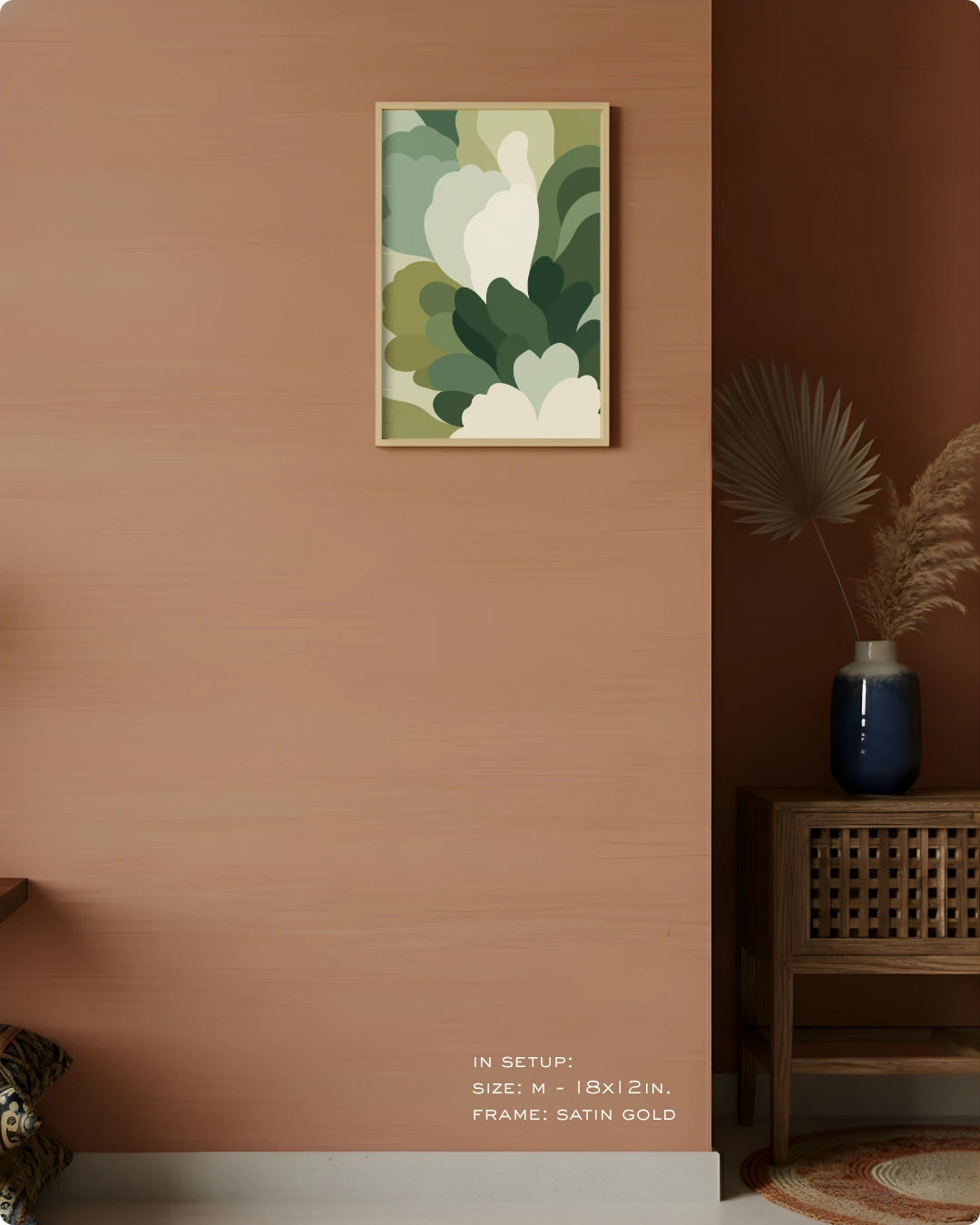 A framed abstract botanical art print with green, beige, and white organic shapes hangs on a warm terracotta wall in a styled interior. A dark wood side table with a blue vase and dried palm leaves sits to the right.