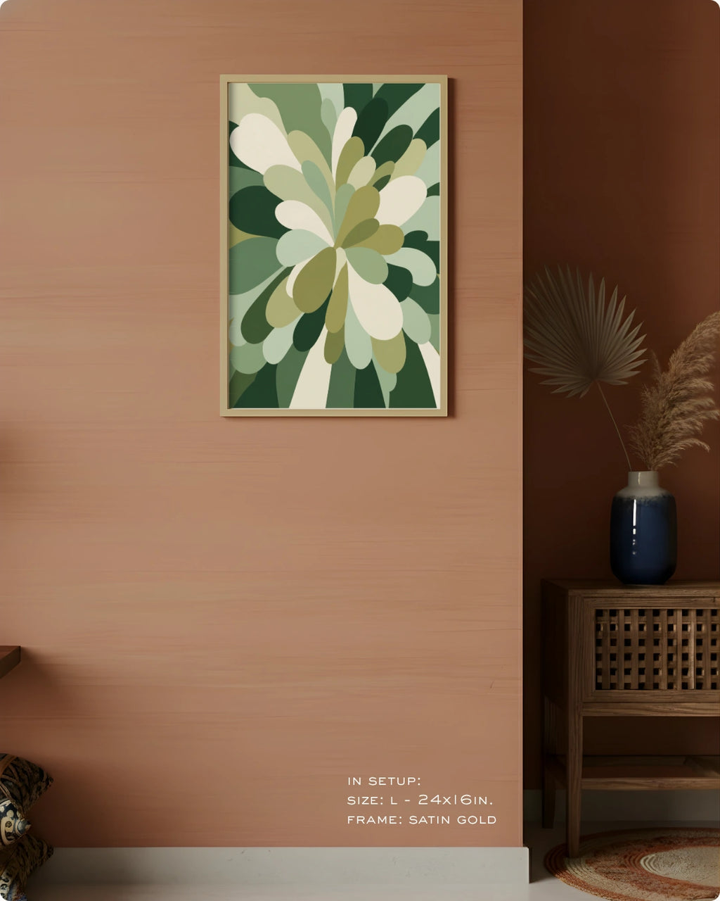 A framed abstract botanical art print with green, beige, and white organic shapes hanging on a warm peach wall in a styled interior. A wooden side table with a blue vase and dried palm leaves sits to the right, and small text at the bottom reads 'IN SETUP: SIZE L - 24KG/IN.