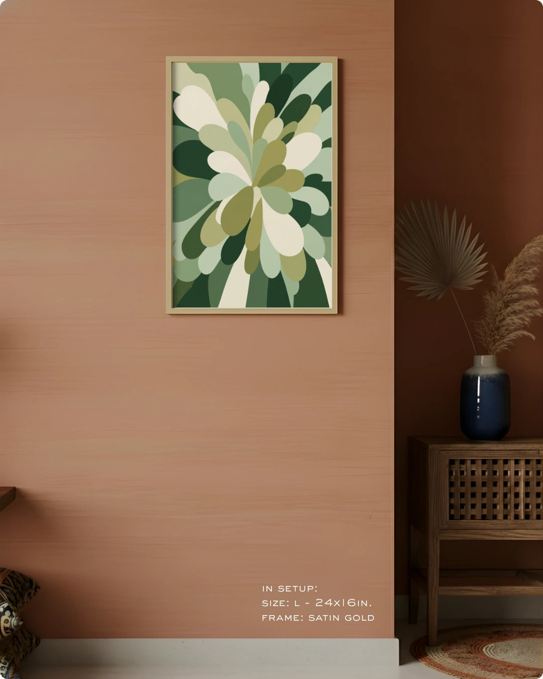 A framed abstract botanical art print with green, beige, and white organic shapes hanging on a warm peach wall in a styled interior. A wooden side table with a blue vase and dried palm leaves sits to the right, and small text at the bottom reads 'IN SETUP: SIZE L - 24KG/IN.