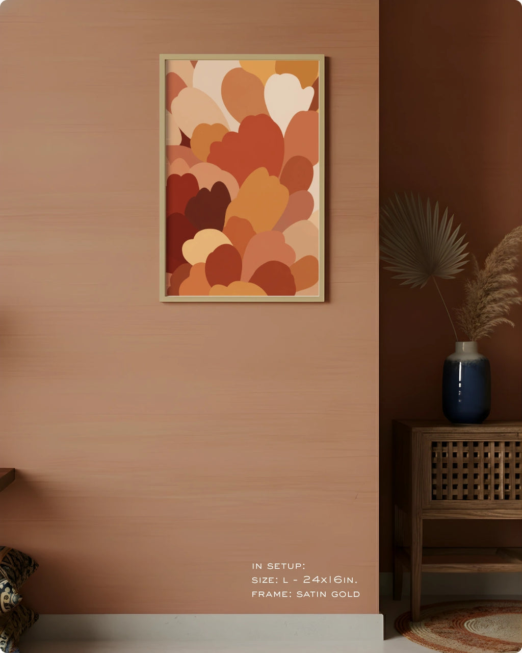 A framed abstract art print featuring overlapping heart-like shapes in warm earthy tones, displayed on a textured peach wall in a styled interior setting with a wooden side table, blue vase, and dried palm leaves.