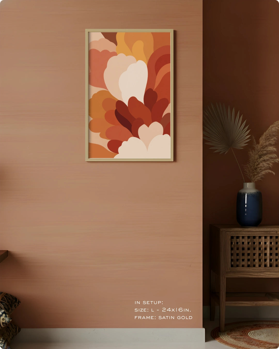 A framed abstract floral art print in warm earthy tones hangs on a peach-colored wall in a styled interior setting with a wooden side table, blue vase, dried palm leaves, and a rug.