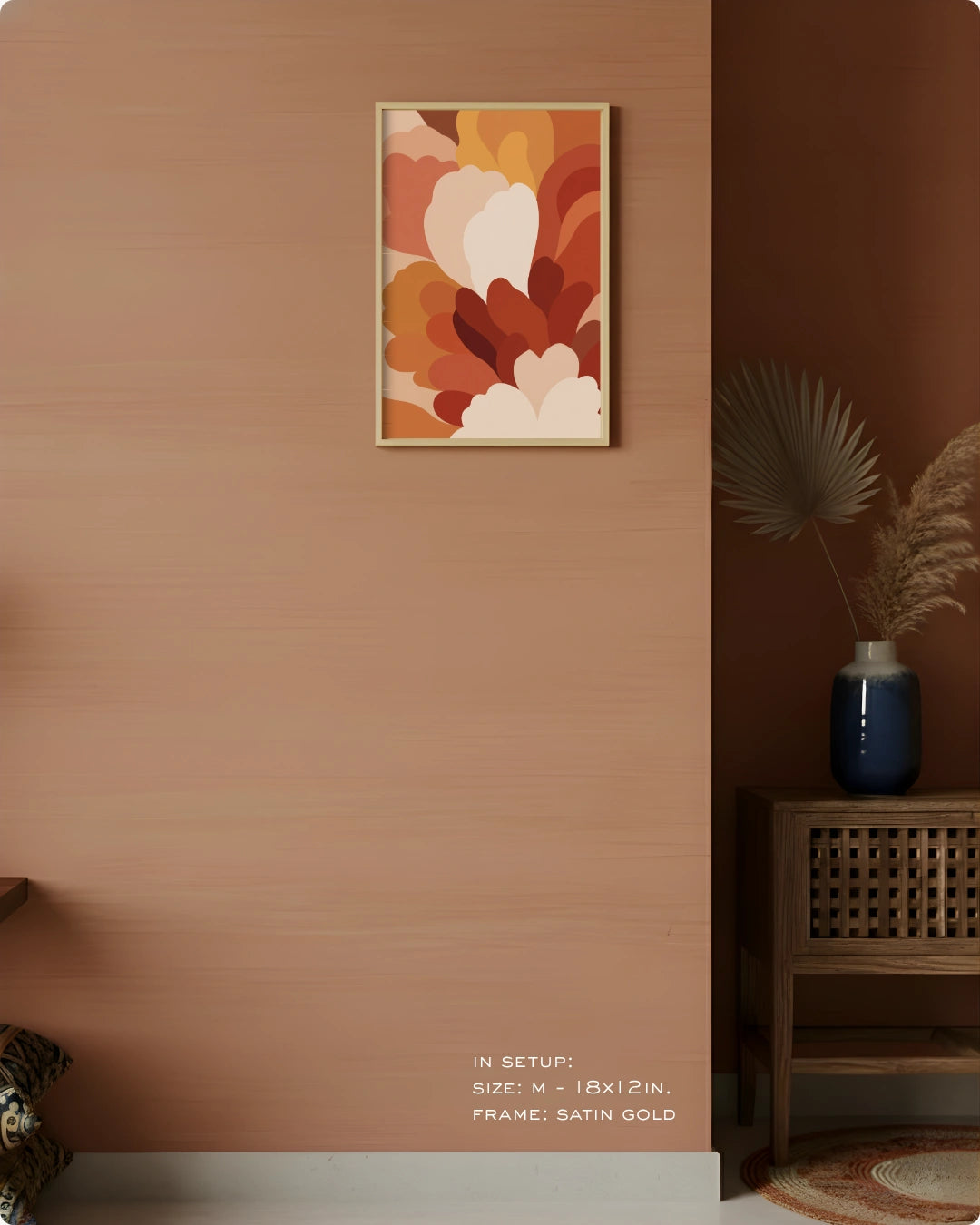 A framed abstract floral wall art print hangs on a warm terracotta wall in a styled interior. The artwork features layered abstract petals in cream, orange, rust, and brown tones, enclosed in a thin satin gold frame.