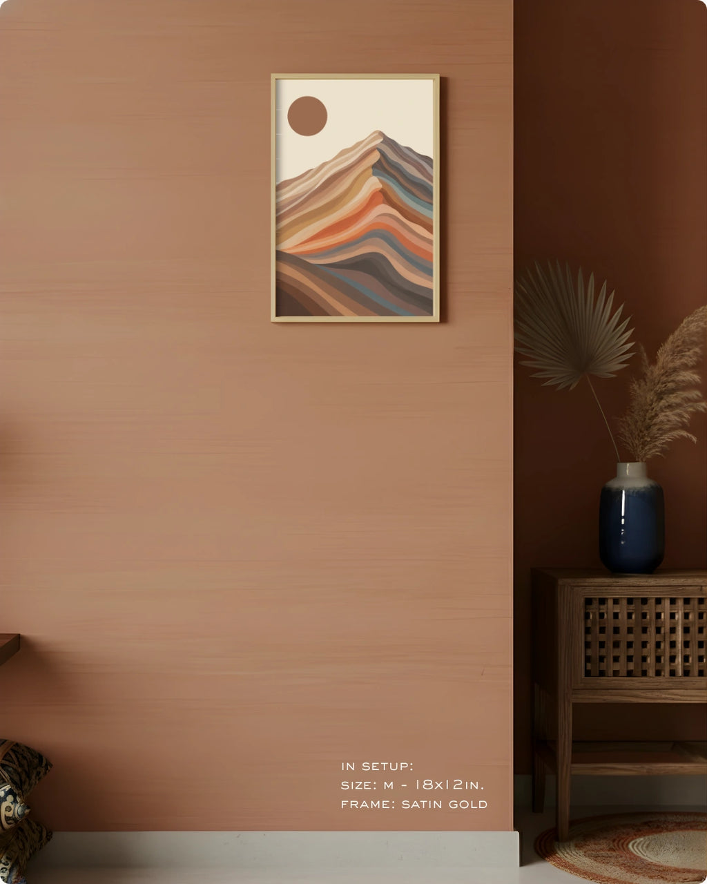 A framed abstract mountain art print with layered warm and cool stripes hangs on a terracotta-colored wall in a styled interior setting. The frame is satin gold.