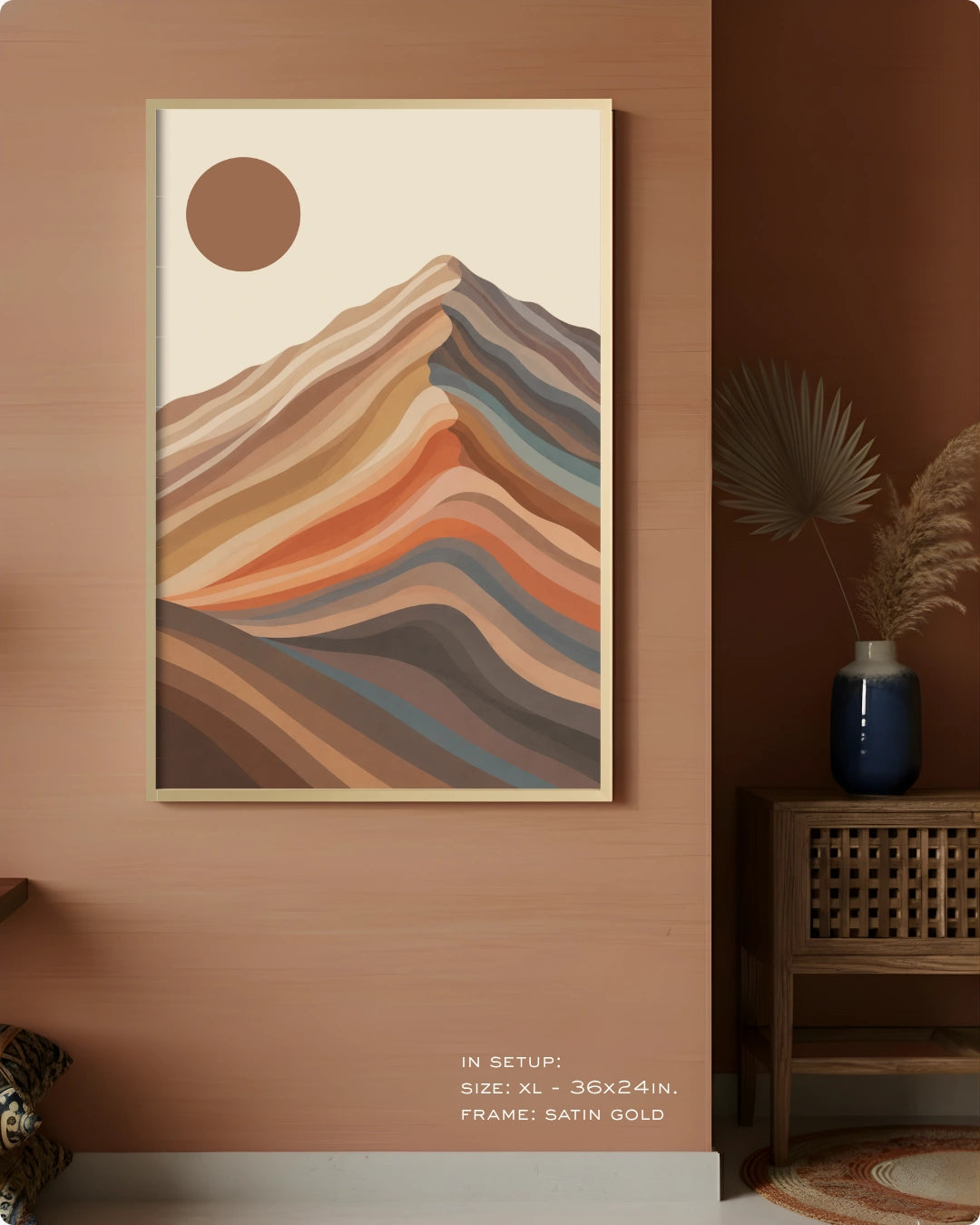 A framed abstract landscape art print featuring layered mountain-like stripes in beige, brown, orange, blue, and gray, hanging on a warm terracotta wall in a styled interior setting.