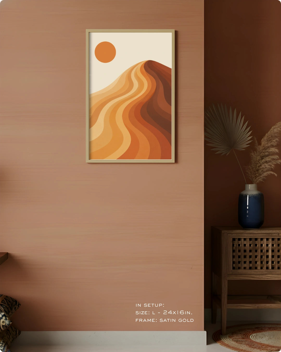 A framed abstract desert landscape wall art print hangs on a warm terracotta wall in a styled interior. The artwork shows layered orange and tan dunes with a sun in the upper left, set in a satin gold frame.