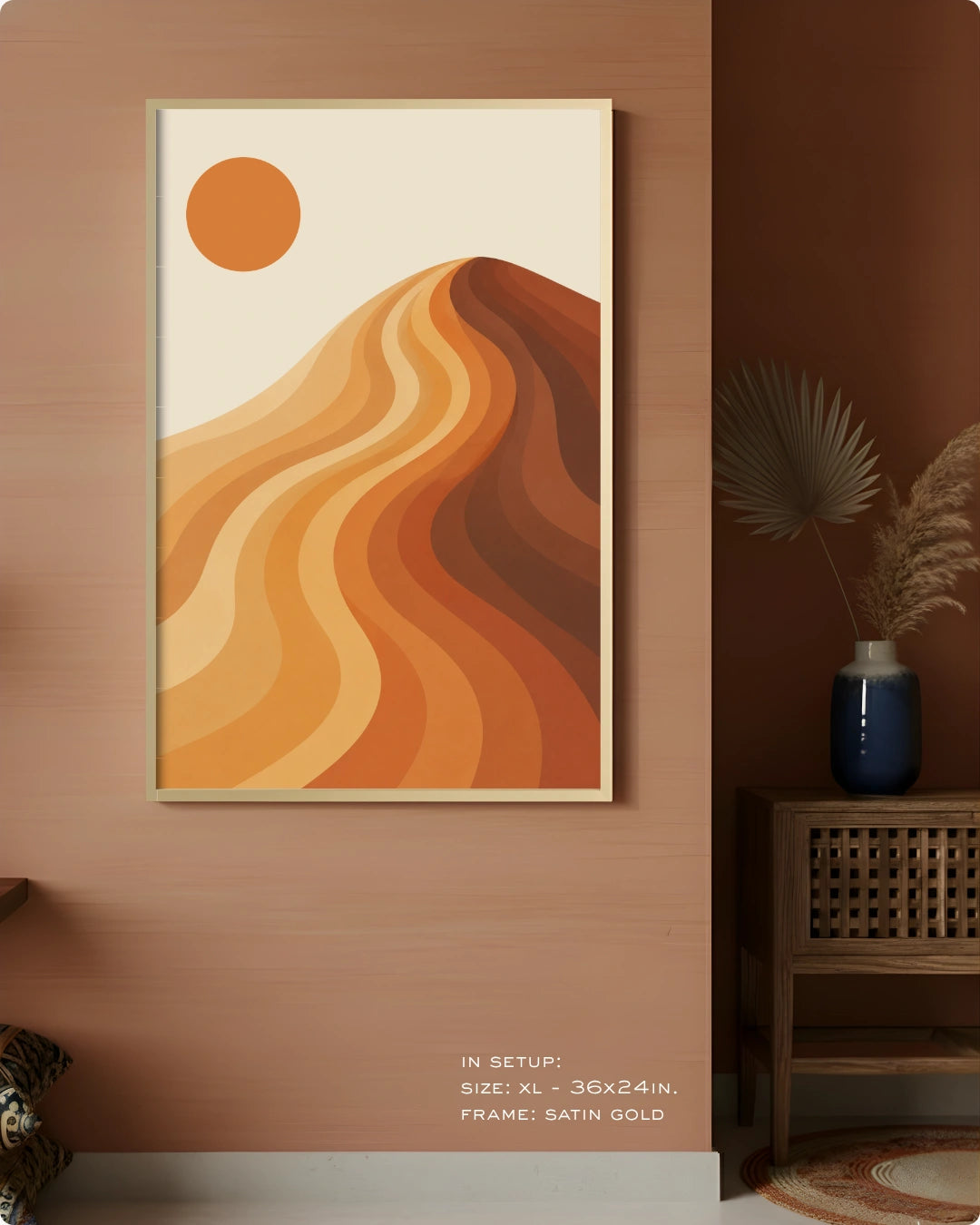 A framed abstract landscape art print featuring wavy orange and brown hills under a solid orange sun on a cream background, displayed on a warm terracotta wall in a styled interior setting.