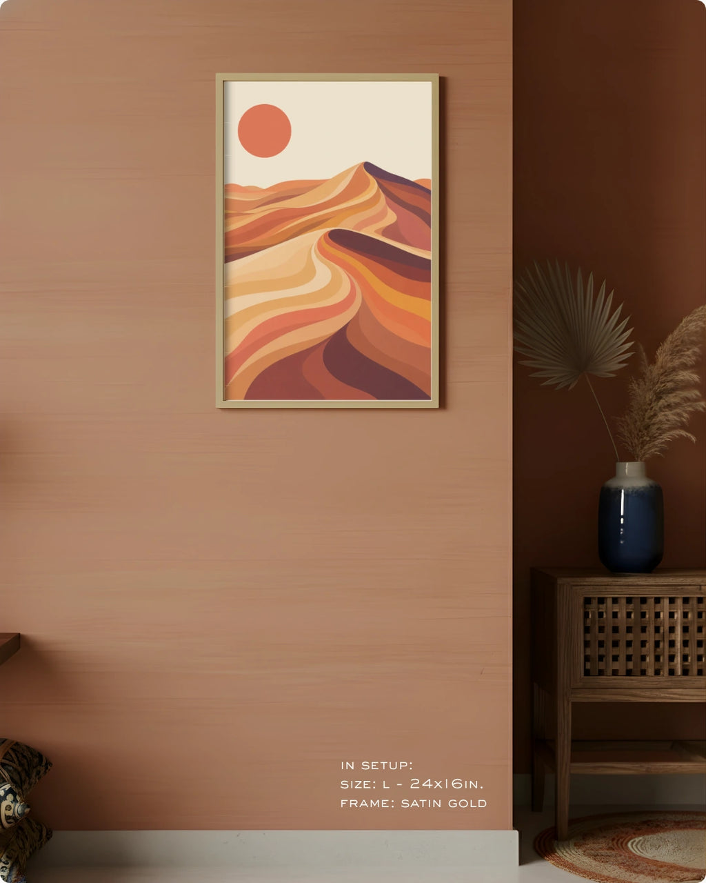 A framed desert landscape art print hangs on a warm terracotta wall in a styled interior. The artwork features layered orange, tan, and brown dunes with a red sun on an off-white background.