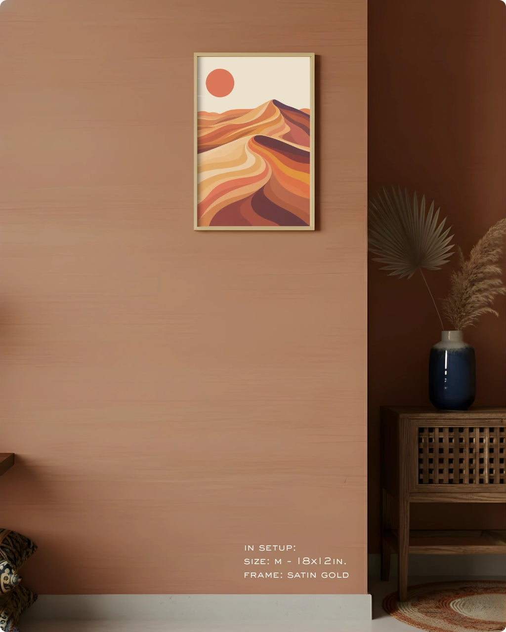 A framed abstract desert landscape art print hangs on a warm terracotta wall in a styled interior. The print features flowing orange and brown dune-like shapes with a small orange sun on a cream background, in a satin gold frame.