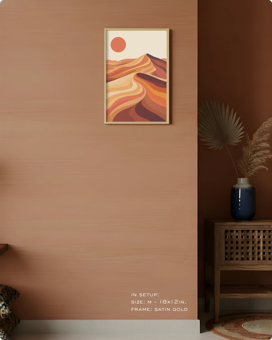 A framed abstract desert landscape art print hangs on a warm terracotta wall in a styled interior. The print features flowing orange and brown dune-like shapes with a small orange sun on a cream background, in a satin gold frame.
