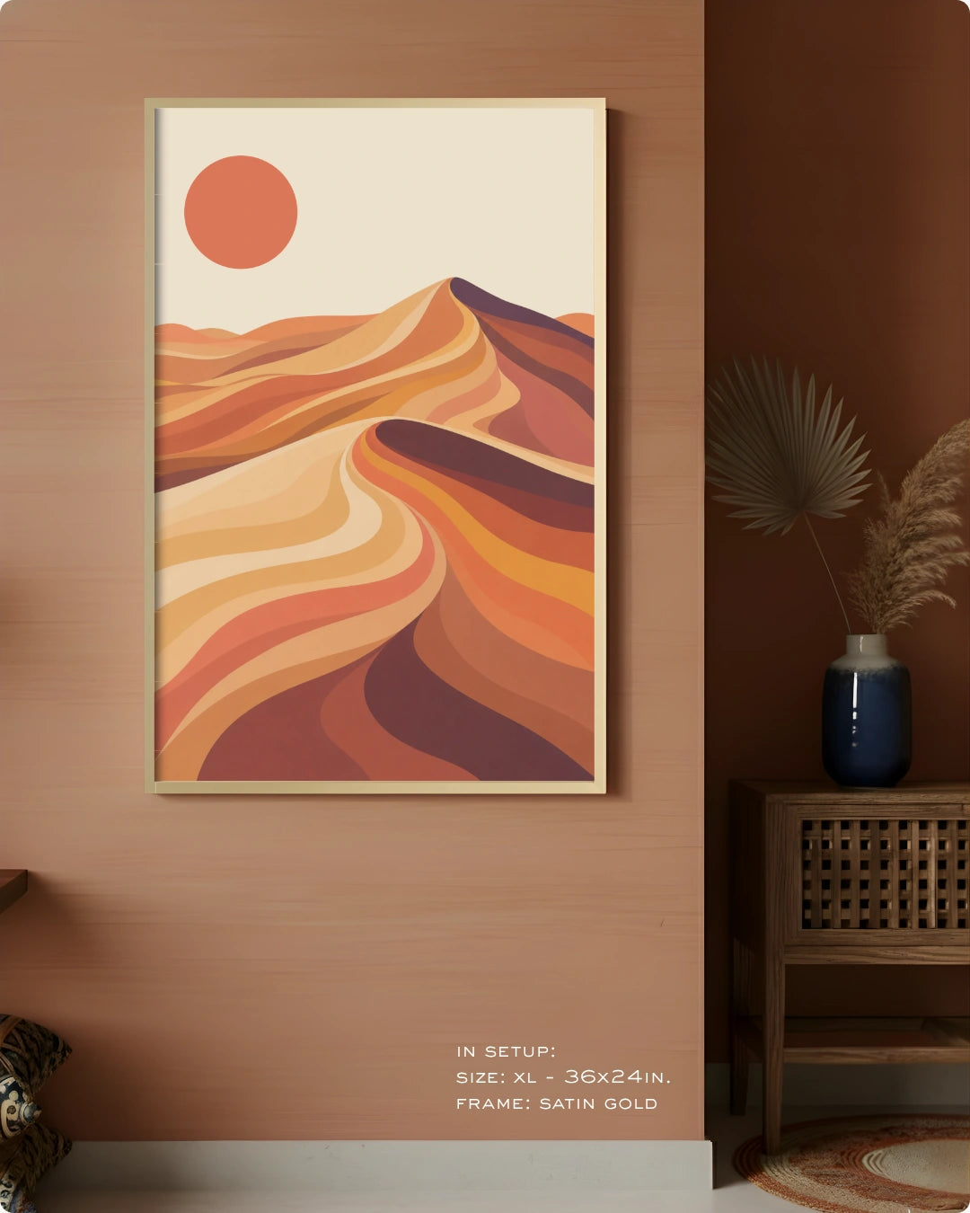 A framed desert landscape art print hangs on a warm brown wall in a styled interior. The artwork shows layered orange, tan, and brown dunes with a large orange sun on a cream background.