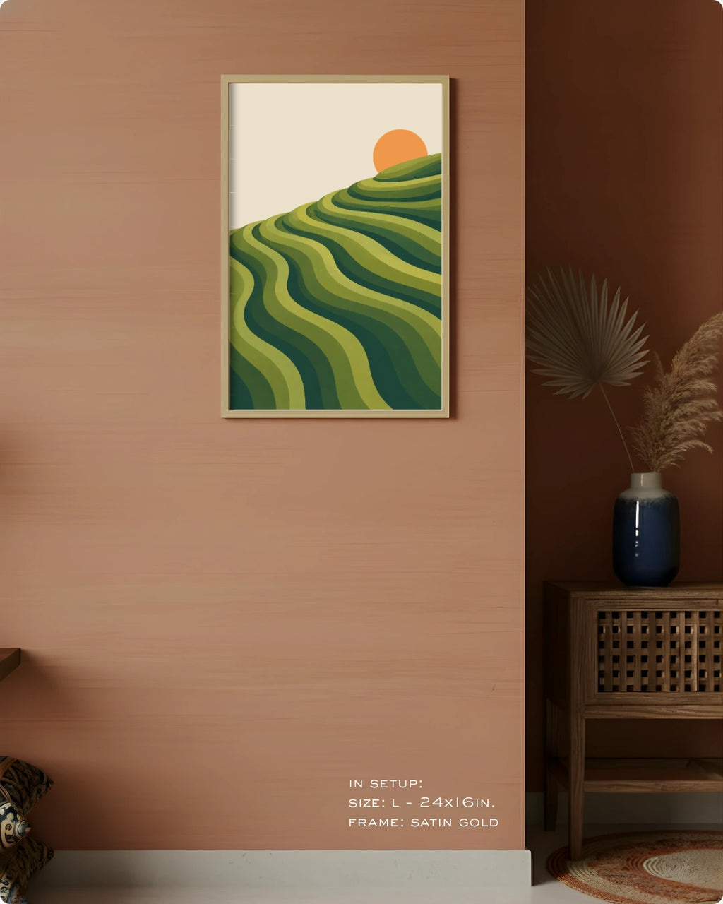 A framed wall art print hangs on a warm terracotta wall in a styled interior. The artwork shows layered green hills with a small orange sun on an off-white background.