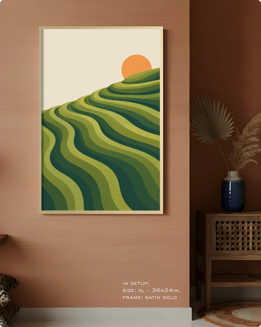 A framed wall art print hangs on a warm terracotta wall in a styled interior. The artwork shows layered green hills with a single orange sun on a cream background.